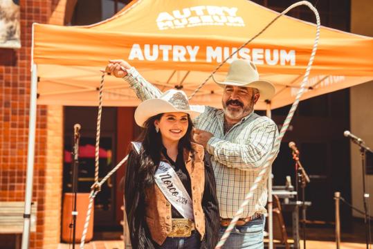 A woman in a cowboy hat and sash smiles as a man in a cowboy hat swings a rope by her. They stand in front of an orange tent with "Autry Museum" written on it. A microphone stand is visible in the background.