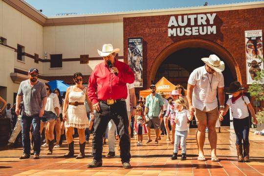 A group of people in cowboy hats and boots are line dancing outside the Autry Museum on a sunny day. The scene is lively, with a mix of adults and children participating. The museum's brick facade is visible in the background.