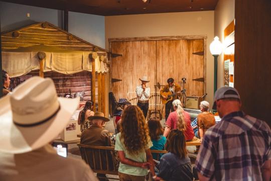 A cozy indoor performance with a man playing a guitar and another speaking or singing, both wearing cowboy hats. An audience of diverse ages watches, seated and standing. The setting includes a rustic backdrop resembling a log cabin.