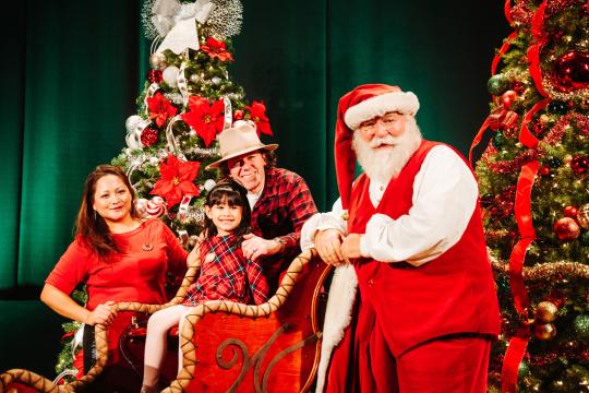 A family and Santa Claus pose together by decorated Christmas trees. The young girl sits in a festive sleigh, smiling, as her parents stand nearby. Santa stands at the front, wearing his classic red suit and hat.
