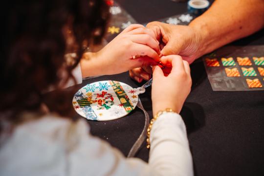 A child and an adult work together to decorate a round ornament with festive stickers, including snowflakes and a reindeer, on a black table. The child's curly hair and bracelet are visible.