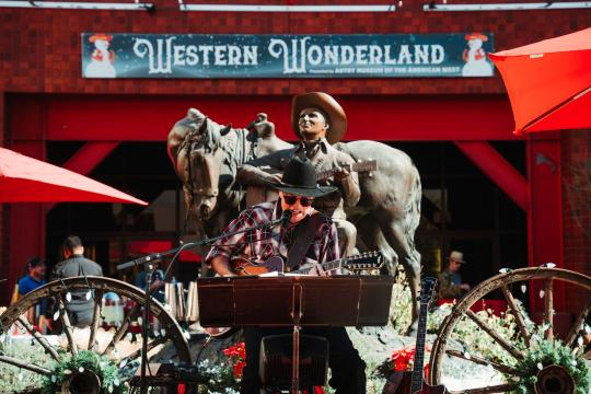 A musician in a cowboy hat performs outdoors in front of a Western-themed statue and a "Western Wonderland" sign, surrounded by festive decorations and red umbrellas.