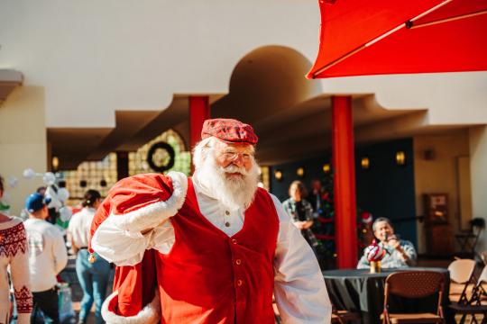 A man dressed as Santa Claus in a red vest and hat carries a red sack over his shoulder, smiling. He stands indoors near tables and people, with festive decorations visible in the background.
