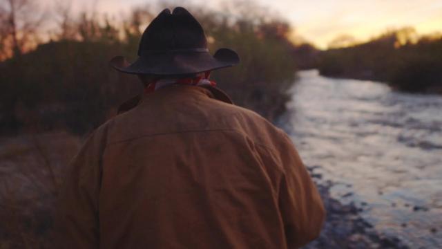 A person wearing a cowboy hat and brown jacket stands near a river at sunset, facing away from the camera with trees and a colorful sky in the background.