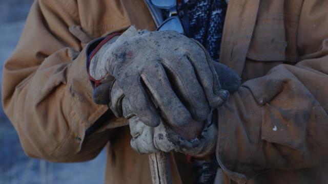 Close-up of a person wearing heavy, worn work gloves and a brown jacket, holding onto a tool or stick, with another gloved hand resting on top. Glasses hang from their neck. Their face is not visible.