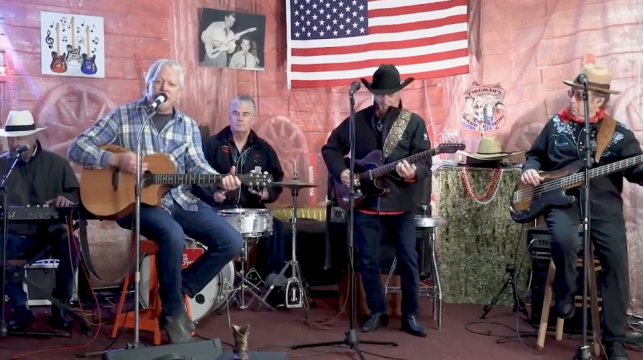 A four-member country band performs indoors; one sings and plays acoustic guitar, one is on drums, one plays electric guitar in a cowboy hat, and one plays pedal steel. An American flag and country decor hang behind them.