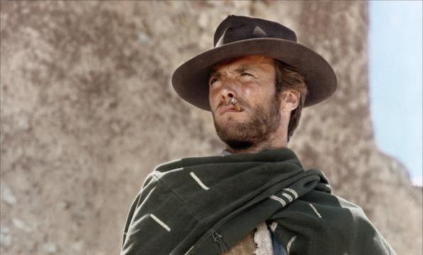 A man wearing a wide-brimmed hat and a poncho stands outdoors, squinting into the distance with a cigarette in his mouth. The background is a sunlit, textured wall.