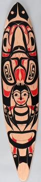 A wooden surfboard decorated with bold black and red Indigenous Northwest Coast art, featuring symmetrical animal and human designs with prominent faces and abstract patterns on a natural wood background.