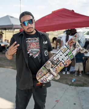 A man wearing sunglasses and a black outfit holds a skateboard with colorful graphics, standing outdoors near red tents. He makes a peace sign with his hand and looks at the camera.