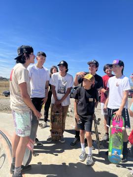 A group of boys with skateboards stand together at a skatepark on a sunny day, smiling and talking. They wear casual clothes and hats, and the sky is clear and blue.