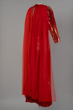 A vibrant red evening gown with sheer, flowing chiffon draped over one shoulder; the dress has shiny, metallic red and gold striped sleeves and hem, creating a dramatic, elegant look against a plain grey background.