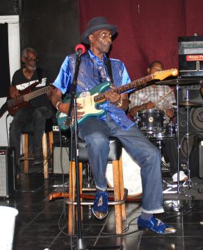 An older musician in a blue shirt, hat, and matching shoes plays electric guitar on stage, seated, with a band including a bassist and drummer behind him. The scene has a casual, intimate club atmosphere.