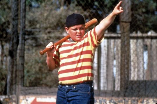A boy in a yellow and red striped shirt, blue jeans, and a backwards cap holds a baseball bat over his shoulder and points into the distance, standing on a baseball field.