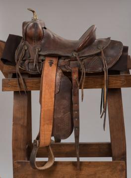 A worn brown leather Western saddle with a horn, stirrups, and saddle strings rests on a wooden saddle stand against a plain gray background.
