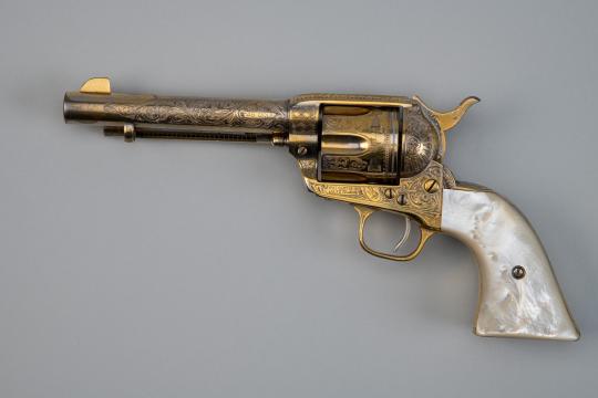 A decorative vintage revolver with intricate engravings on the barrel and cylinder, featuring a gold-toned frame and a shiny mother-of-pearl grip, displayed on a plain gray background.