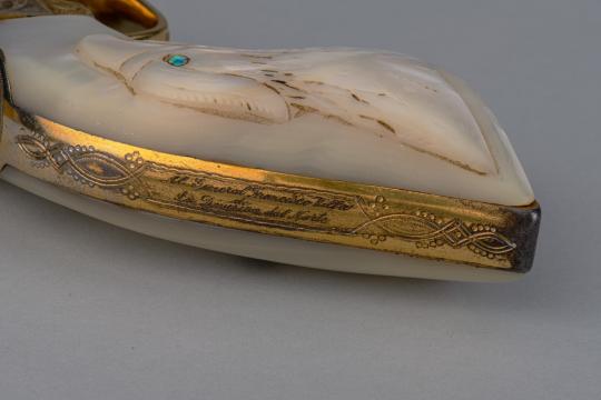 A close-up of an ornate handle, possibly from a dagger or sword, with intricate gold detailing and engraved text in Spanish. The handle is cream-colored with a carved pattern and a small green gem.