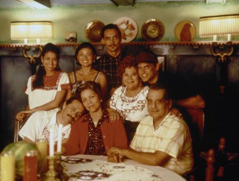 A group of nine people, including men and women of varying ages, smile and pose together around a dining table decorated with candles and a lace tablecloth in a warmly lit, homey room.