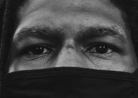 black and white closeup photo of a Black individual with dreadlocks and a mask covering the lower half of their face, leaving their eyes visible to look at the camera