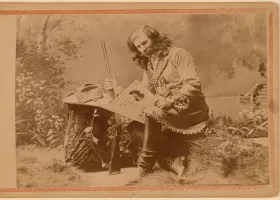 A vintage sepia photograph of a person in frontier attire sitting on a tree stump. They hold a rifle and a book while wearing a long coat with fringe details. The background features a painted outdoor scene with trees and foliage.