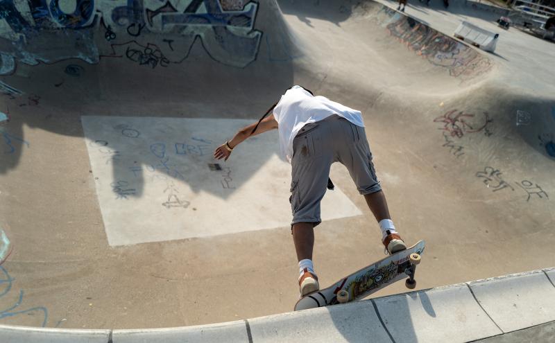 A skateboarder in a white shirt and gray shorts prepares to drop into a concrete skatepark bowl covered in graffiti and drawings, captured from behind under bright daylight.