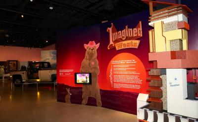 A museum exhibit titled "Imagined Wests" features a large display wall with a bear wearing a cowboy hat and a rabbit. A small truck is visible in the background, and the foreground has a model structure labeled "Los Pobladores." The setting is well-lit and spacious.