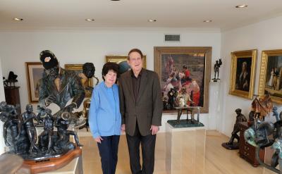 An older couple stands smiling in an art-filled room. They are surrounded by various sculptures and framed paintings on the walls. The sculptures include human figures and animals, displayed on pedestals and shelves. The room is well-lit.