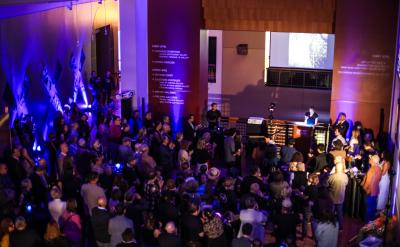 A large crowd gathers in a dimly lit room with blue lighting. People are standing and mingling, facing a stage where speakers are presenting. Text is displayed on the wall behind the stage.