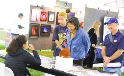 A woman and two teenagers smile and interact with an artist seated at a table at an art fair, while another man stands beside them holding a rolled poster. Colorful artwork is displayed on panels in the background.