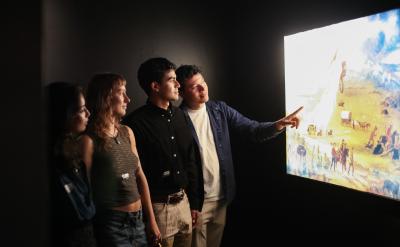 Four people stand in a dark room closely observing and discussing a brightly lit display or painting on the wall, with one person pointing at the artwork.