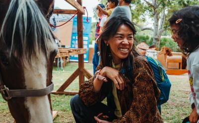 A woman crouches and smiles at a young girl near a horse at an outdoor event, with trees and other people visible in the background.