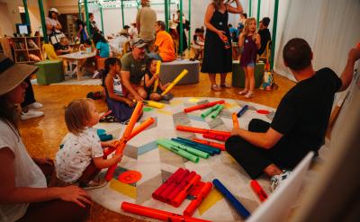 Children and adults sit on a rug in a brightly lit playroom, playing with colorful plastic tubes. Other groups are engaged in activities in the background, creating a lively, interactive environment.