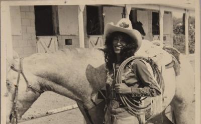 A smiling woman in cowboy attire, including a hat and fringed chaps, stands next to a saddled horse outside a stable, holding a coiled rope. The photo is in black and white.