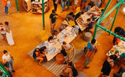 Overhead view of a busy indoor event with people of all ages gathered around tables covered in art supplies and crafts, socializing, and participating in creative activities under bright lighting.