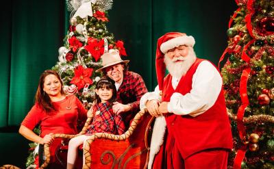 A family poses with Santa Claus in front of decorated Christmas trees. The child sits in a sleigh, smiling, while Santa and her parents stand nearby, all dressed in festive holiday attire.
