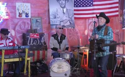 Three musicians perform on stage in a country-themed setting with guitars, American flags, and photos of country legends on the wall. One plays keyboard, one is on drums, and the third plays guitar and sings wearing a cowboy hat.