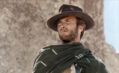 A man wearing a wide-brimmed hat and a poncho stands outdoors, squinting into the distance with a cigarette in his mouth. The background is a sunlit, textured wall.