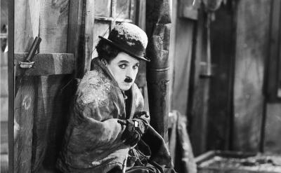 A person dressed in old-fashioned clothes and a bowler hat, with a small mustache, sits shivering in a rustic, snowy cabin, clutching their coat tightly for warmth.