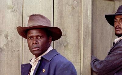Two men in old-fashioned clothing and wide-brimmed hats stand in front of a wooden wall; one faces forward while the other leans against a door, looking sideways.