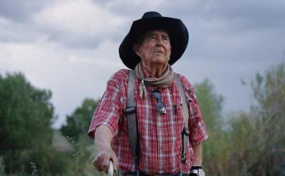 An elderly man wearing a black hat, red plaid shirt, and suspenders stands outdoors with greenery and cloudy skies in the background, looking thoughtfully into the distance.