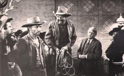 A group of men in cowboy hats and Western attire gather indoors; one man holds a coiled rope, another appears to be loading a gun, and an older man sits in the background, all in a tense black-and-white scene.