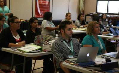 A group of adults sits at tables in a classroom, attentively listening and taking notes. Some have laptops open, and papers and binders are on the tables. The room is well-lit with windows in the background.