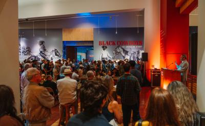 A crowd listens to a speaker at a podium in a gallery space with cowboy-themed artwork and a large sign reading “Black Cowboys: An American Story” on the wall in the background. The room is warmly lit with orange and blue lights.