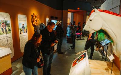 People view exhibits in a museum, including a large white horse sculpture and informational panels on the walls. The room is warmly lit and several visitors are reading displays or interacting with exhibits.