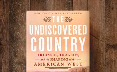 Book titled "The Undiscovered Country" by Paul Andrew Hutton on a wooden surface. The cover shows a group of people on horseback in a western landscape at sunset.