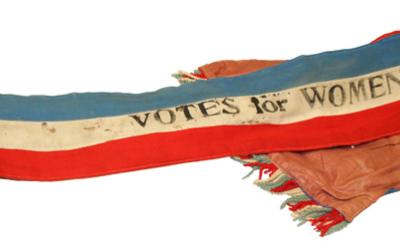 A fabric sash with blue, white, and red stripes, featuring the text "VOTES for WOMEN" printed in black. The ends have multicolored fringe and appear slightly worn.