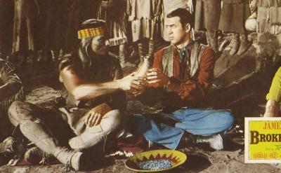 Three men sit on the ground in traditional Native American and Western attire. One man hands a peace pipe to another. A yellow "Broken Arrow" movie poster with James Stewart's name appears in the lower right corner.