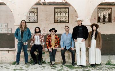 Six people stand in front of a weathered building with arched windows. They wear casual and bohemian-style clothing, including hats, sunglasses, and patterned shirts. The mood appears relaxed and vintage.