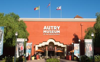 The front entrance of the Autry Museum of the American West, with banners and three flags atop the building. Tall trees and clear blue sky are visible around the museum.