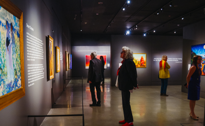 People view colorful paintings on the walls of a dimly lit art gallery. The space has polished floors, spotlights illuminating the artwork, and visitors standing and reading descriptions or admiring the art.