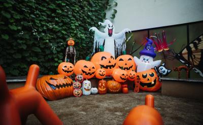 A group of inflatable Halloween decorations, mostly orange pumpkins with carved faces, a large white ghost, and a witch, are arranged outdoors near a wall covered in green vines.